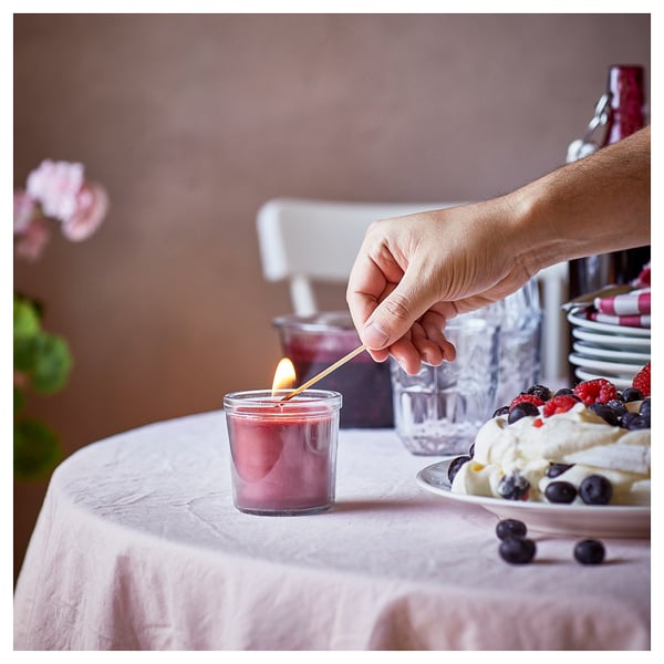 Рука зажигает маленькую красную свечу STÖRTSKÖN на столе, рядом с десертом, бокалами и цветами.