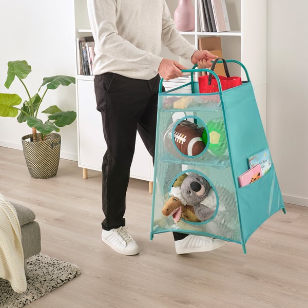 Человек собирает игрушки в корзину для хранения из синей сетки с карманами и ручками. Бункер имеет круглые отверстия для легкого доступа.
