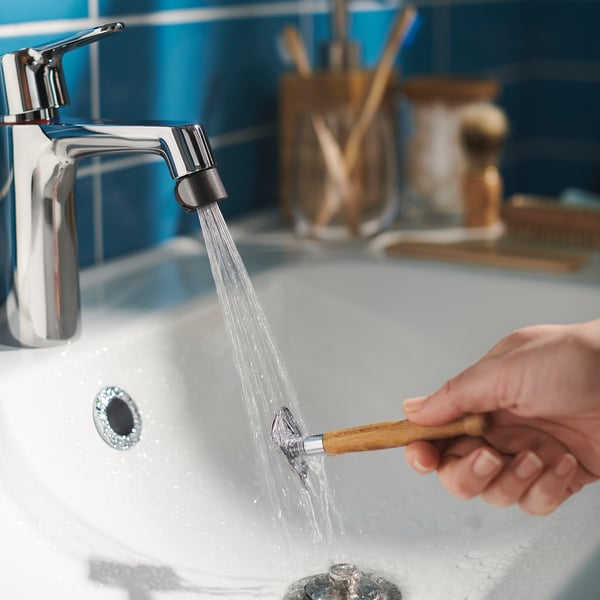 Hand holds wooden ÅBÄCKEN nozzle under running water from chrome tap, sink.