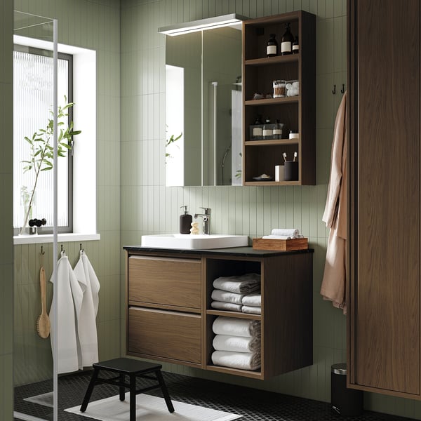 Modern green bathroom with wall-mounted brown oak cabinet, white sink, open shelves, and hanging towels. Mirror and plant by window.