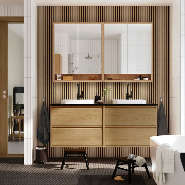 Modern bathroom with light wood ÄNGSJÖN vanity, dual sinks, wooden backdrop, large mirrors, and towel racks.