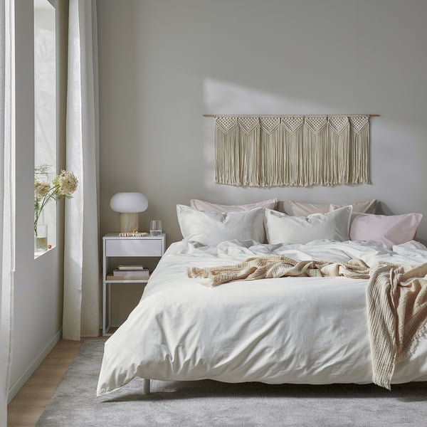 Modern bedroom with white ÄNGSLILJA duvet cover and pillowcases on a large bed. Wall hangings, lamps, and flowers decorate the space.