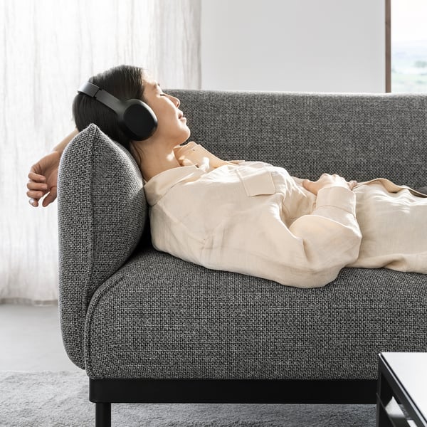 A person reclines on a grey ÄPPLARYD sofa, listening to headphones, comforted by soft beige blankets in a cosy living room.