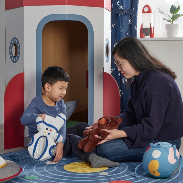 Person & boy play with toys in space-themed room. The person holds a bear; the person holds a space bear. A rocket playset is nearby.