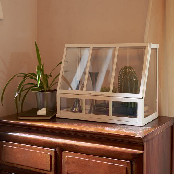 A white wooden plant greenhouse sits atop a dresser, displaying small plants and decorative vases.
