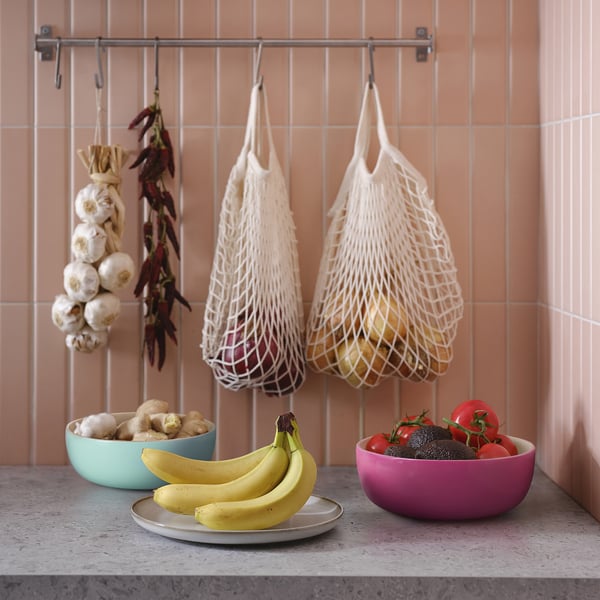 Kitchen counter with fresh produce. Two colourful bowls: teal, magenta, and silver. Net bags holding onions. Peach tiled wall.