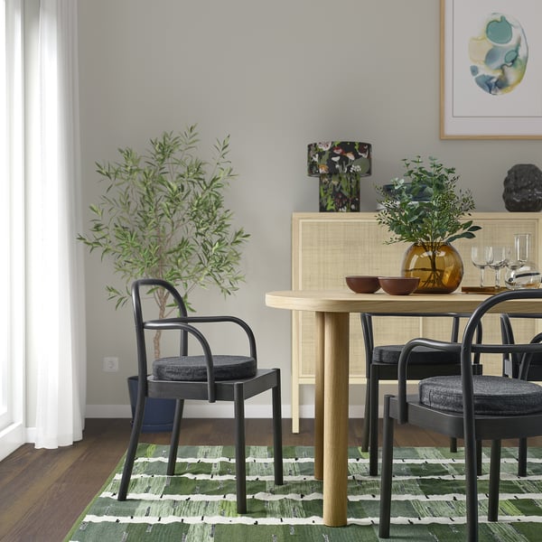 Modern dining area with round light wood table, four sleek black ÅKERVINDEFLY chair pads, green striped rug, potted plants, glassware, and wall art.