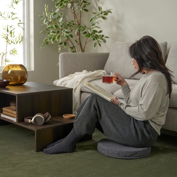 A person sits comfortably on the floor, sipping tea and reading a book on a grey memory foam chair pad that stays in place.
