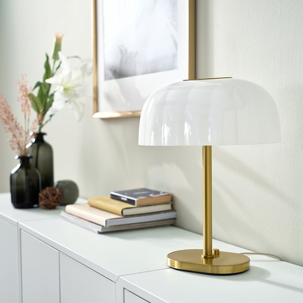 AKTERSPRING lamp on white surface, brass base, white shade. Books, vase, picture nearby.