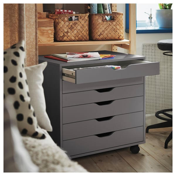 Grey ALEX storage unit with five drawers, one open showing files and supplies. In room with wooden shelves and polka dot pillow.