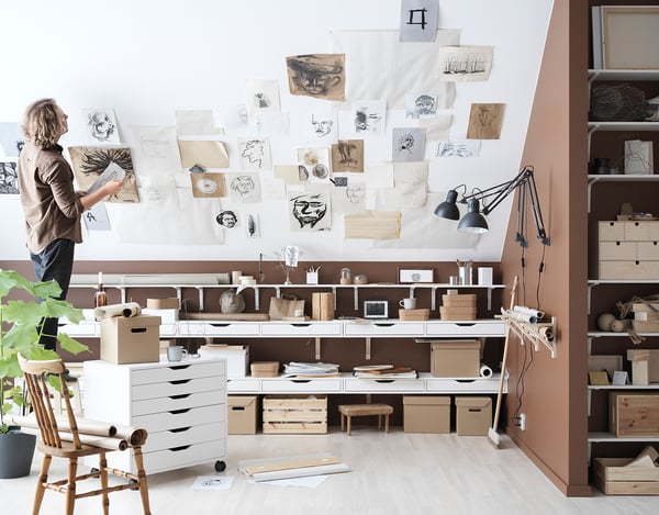Artist in studio with sketches, supplies, ALEX units, chair, plant, lamp, wall drawings.