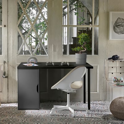 Small, minimalist desk named ALEX, black with a white chair, positioned under large windows, surrounded by plants and decor in a bright room.