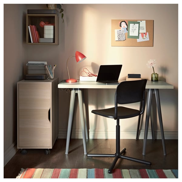 Modern home office with beige ALEX storage, wooden desk, black chair, red lamp, laptop, and colourful rug.