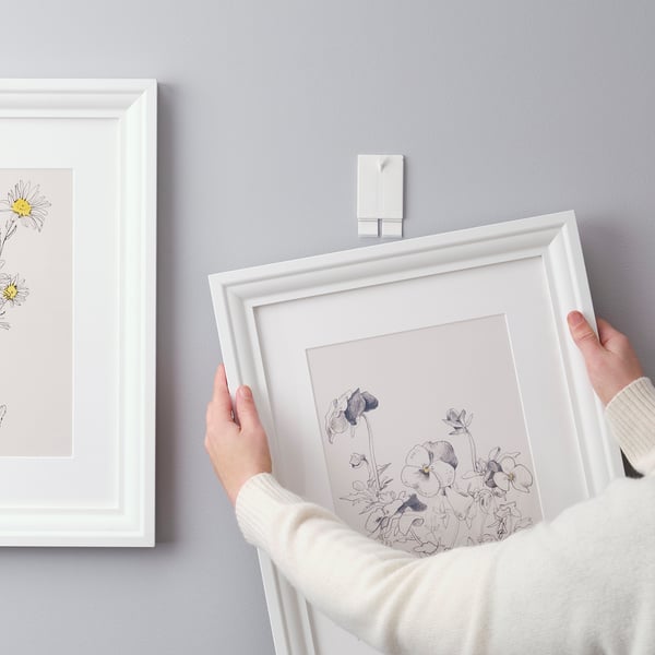 Person holding white frame with floral sketch. Demonstrates ALFTA adhesive hook usage for wall mounting.