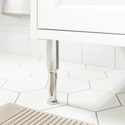 A sleek, stainless steel leg supports a white cabinet on a tiled floor, showcasing modern bathroom furniture.