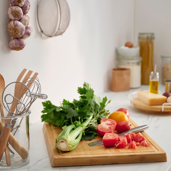 Kitchen scene with APTITLIG bamboo chopping board, featuring fresh veggies, utensils, and hanging garlic. Clean, natural, and efficient.