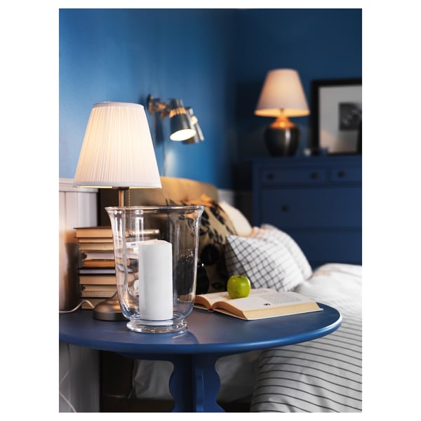 Bedside table withlit lamp, books, apple, striped sheets, blue furniture.