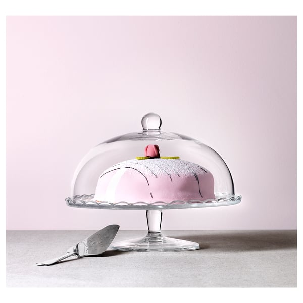 Glass cake stand with clear dome lid, holding pink cake. Silver serving spatula lies beside it.
