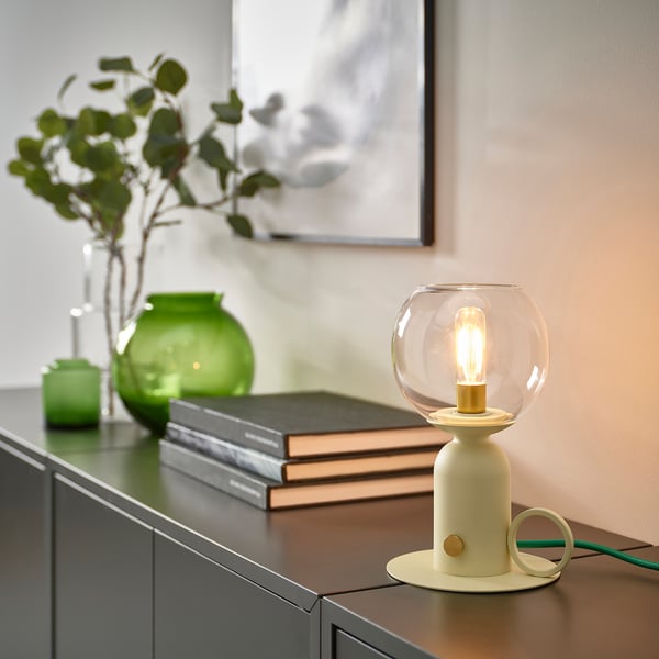 White ÅSKMULLER lamp with glass globe on dark wood, green decor, warm glow.