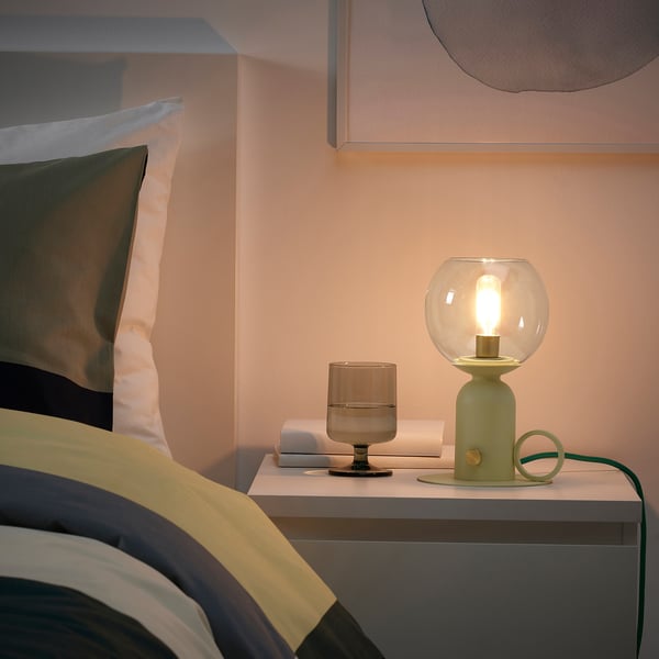 Cosy bedroom with ÅSKMULLER lamp on bedside table. Small, light green, with warm, relaxing light.
