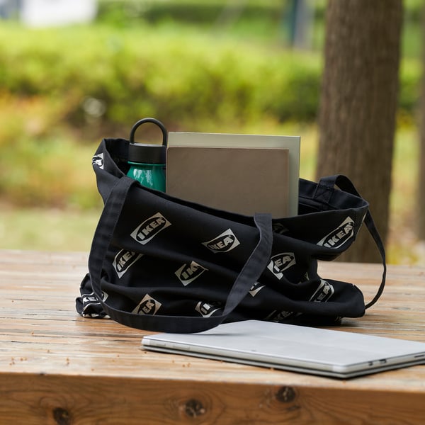 Black ikea tote bag with green water bottle and box on wooden table. Bag has ikea logos and inner pockets. Laptop partially visible.