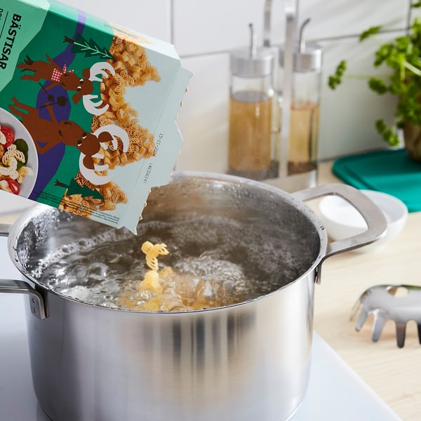 Silver pan boiling pasta with elk-shaped BÄSTISAR packaging being poured in. Kitchen background.