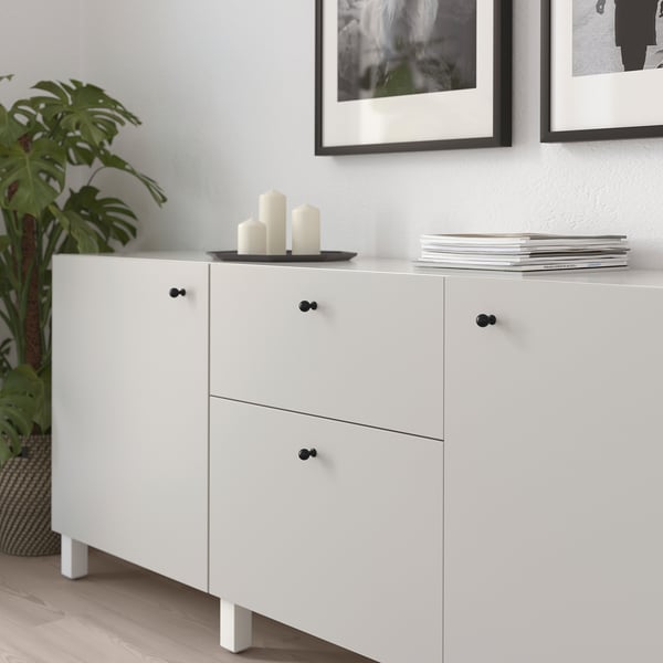 White cabinet with black handles, two drawers, candles, and stack of books. Plant left, framed photos above.