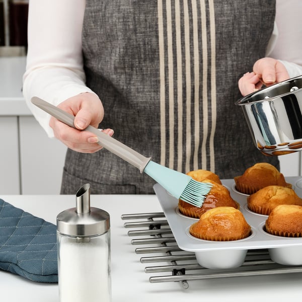 A person brushes baked goods with a silicone brush, ensuring even coverage. A muffin tray and sugar dispenser are nearby.