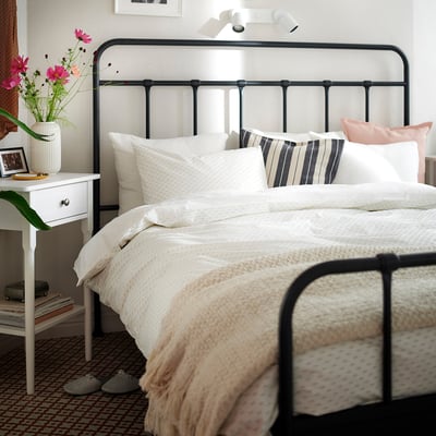 Cosy bed with green/white duvet, pillows, furry blanket, bedside table with books and pink flowers.