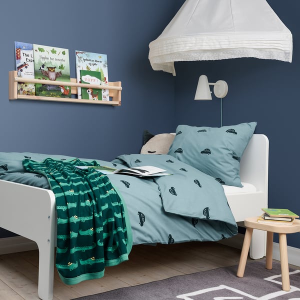 A BARNDRÖM green duvet cover with car prints, made of soft cotton, on a white bed. The wall shelf with books is visible above it.