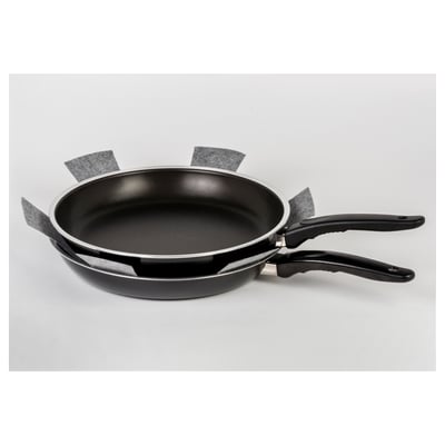 Stacked black frying pans with protective grey fabric strips, displayed on a light surface.
