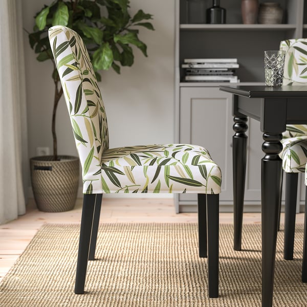 A chair with a green leaf pattern cover and dark wood legs sits near a dining table with a glass and a plant.