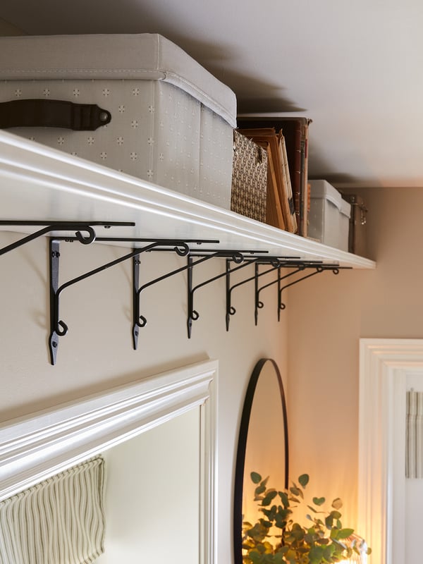 White wall shelf with black metal brackets holds storage boxes and files. Underneath, a round mirror reflects a green plant in a warm room.