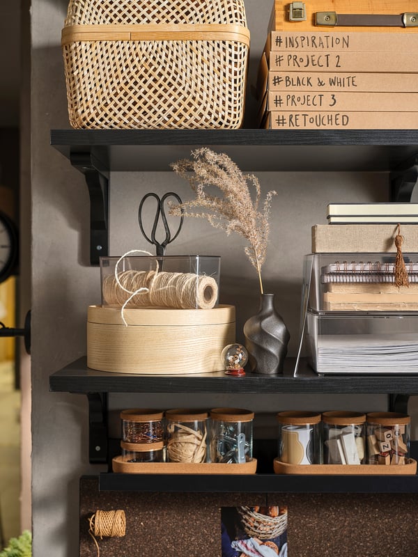 Dark brown wooden shelves holding storage boxes, twine, scissors, and vases against a grey wall.