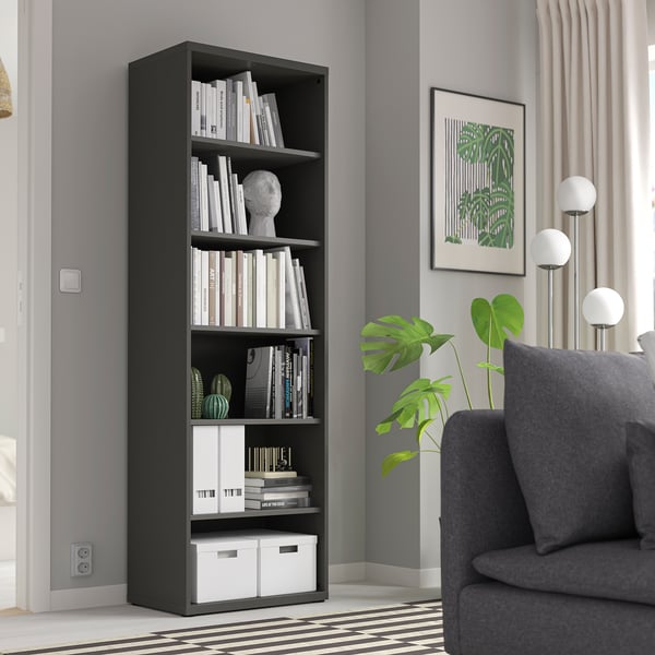 Black BESTÅ bookshelf with books & decor against grey wall. Grey armchair, floor lamps, green plant, and leaf picture frame complete the modern setup.