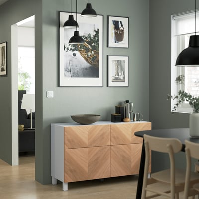 Modern living room with BESTÅ sideboard, drawers, and decor. Black lamps and art on blue-green walls. Wooden chair and dining table in foreground.
