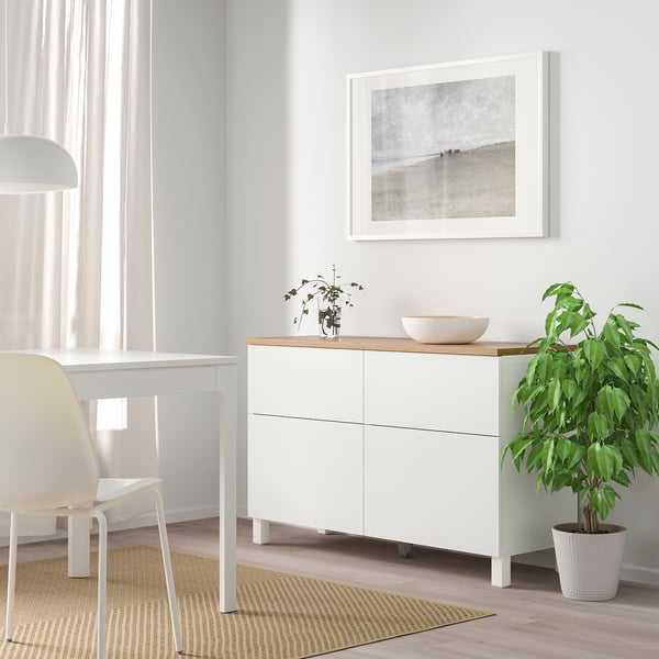 Modern living room with white BESTÅ sideboard, tall white chair, plant in corner, beige rug, and framed artwork.