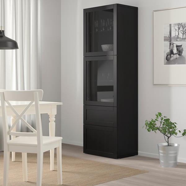 Black BESTÅ cabinet with glass doors, holding wine glasses and bowls, beside a white chair and plant.