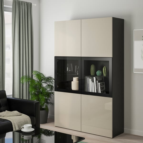 Modern BESTÅ storage cabinet in beige & black, with decorative items behind glass doors, surrounded by a chair, plant, and coffee table.