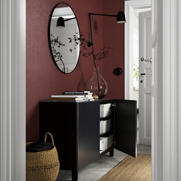 Black sideboard with open doors, storage boxes, maroon wall, mirror, lamps, basket, white door.