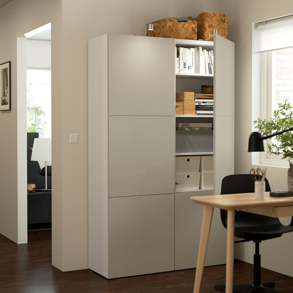 Modern home office with beige BESTÅ storage unit. Shelves adjustable, door open showing organised items. Desk with chair, lamp, and potted plant.