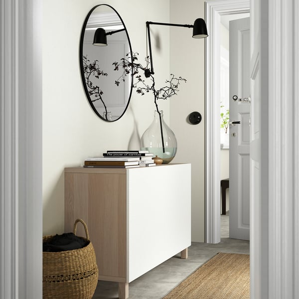 Modern entryway setup with BESTÅ sideboard, a mirror, and a vase with branches. The sideboard has adjustable shelves and is secured to the wall.