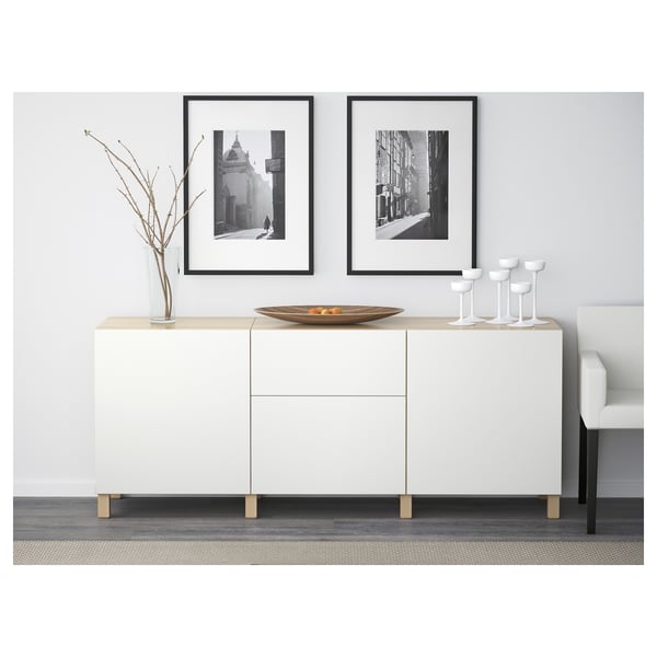 White BESTÅ sideboard with drawers and shelves. Two framed photos hang above it. A vase with branches and a decorative bowl are on top.
