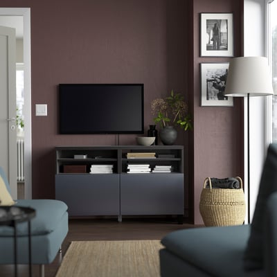 Grey BESTÅ tv bench with tv, books, and drawers, between blue chairs, with lamp and basket.