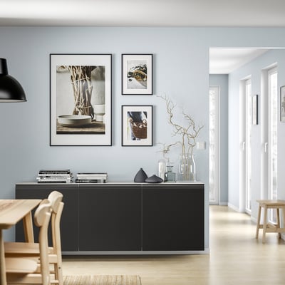 Modern living room a black BESTÅ cabinet with wooden chairs and decor. Three framed photos hang above; a light fixture by the wall.