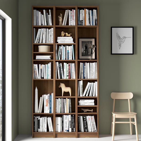 Large wooden BILLY bookcase filled with books, decorated with small figurines, against green wall. Mild wood chair beside it, hummingbird art above.