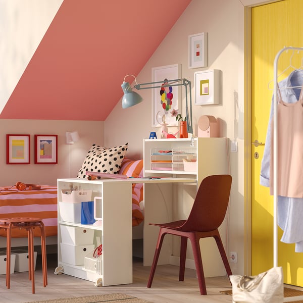 Small home office with pink walls, BILLY white desk & chair, red lamp, shelves with decor, yellow door, clothes rack.