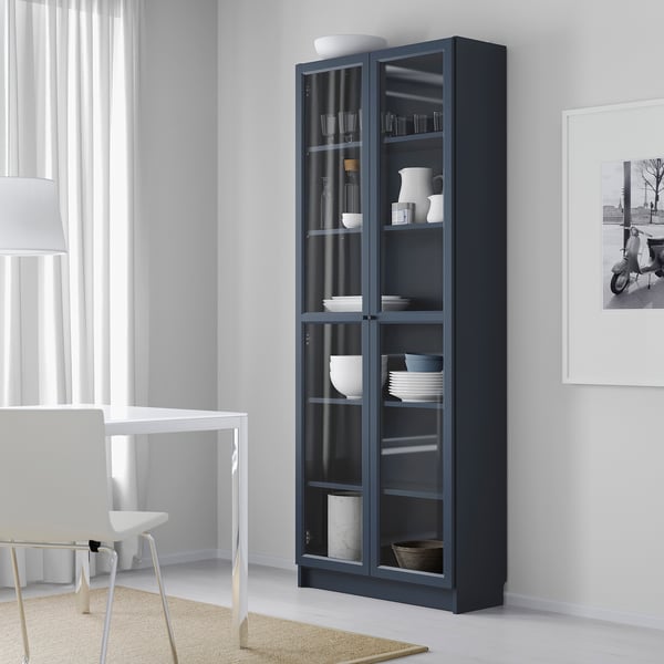 Blue BILLY bookcase with glass doors beside table and chair in minimalist room, displaying white dishes.