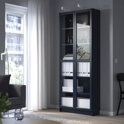 Black BILLY bookcase with glass doors, between a window and chair.