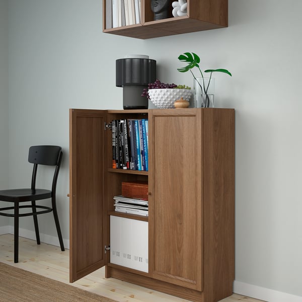 Wooden cabinet with open doors showing books and a shelf with white paper. Top has lamp and decorations.